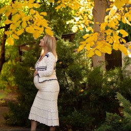 Niebiańska Fota - Kobieta w ciąży w kremowej sukience pozuje w ogrodzie pod drzewem z żółtymi liśćmi. Profil, delikatny uśmiech, naturalne światło.