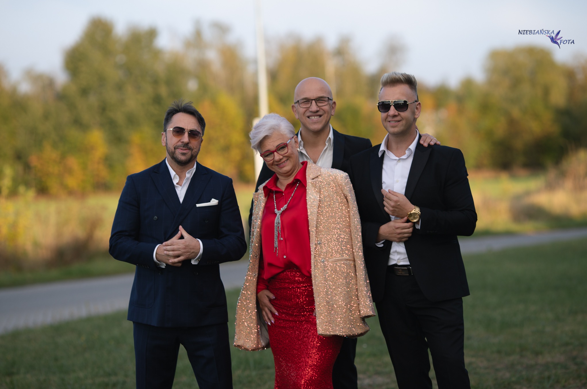Grupowe, plenerowe zdjęcie czterech osób w eleganckich strojach, pozujących na tle zieleni i drzew. Mężczyźni w garniturach, kobieta w czerwonej sukience i cekinowej marynarce.
