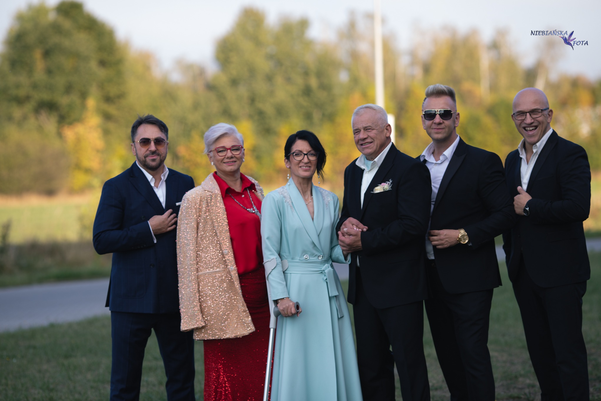 Elegancka sesja grupowa na tle zieleni: trzy kobiety w kreacjach i trzech mężczyzn w garniturach, uśmiechnięci, w naturalnym oświetleniu plenerowym.