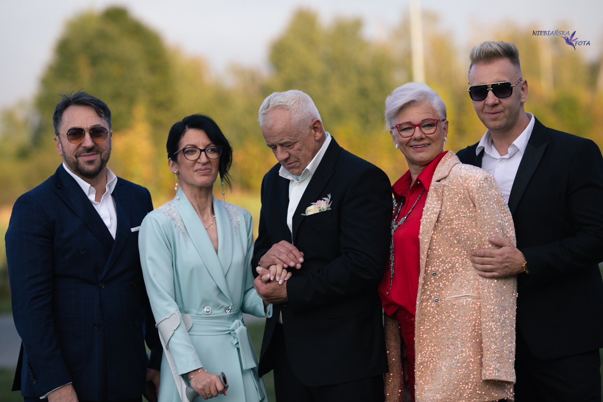 Elegancka, rodzinna fotografia plenerowa z pięcioma osobami w odświętnych strojach, złączone dłonie pary w centrum kadru, zieleń w tle i logo fotografa w prawym górnym rogu.