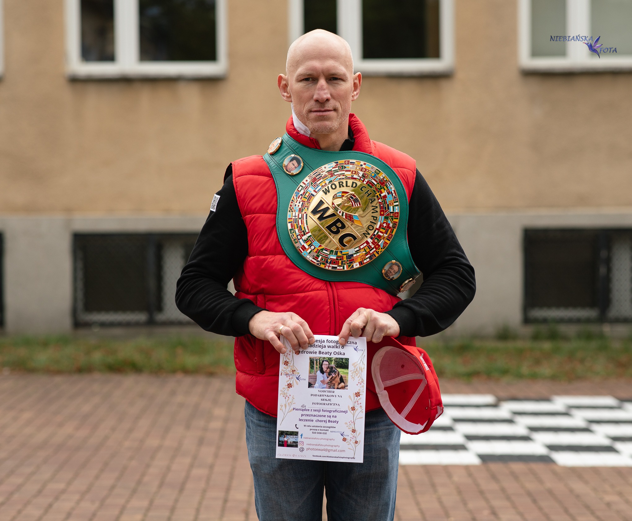 Mężczyzna z pasem mistrzowskim WBC trzyma ulotkę o sesji fotograficznej na leczenie chorej Beaty Ośka. W tle budynek z oknami i logo fotografa.