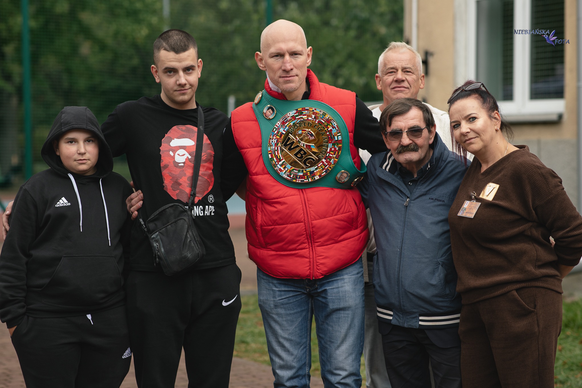 Portret grupowy z mistrzowskim pasem WBC, na tle zieleni i budynku. Uczestnicy różnej płci i wieku, w swobodnych ubraniach. Zdjęcie w plenerze.