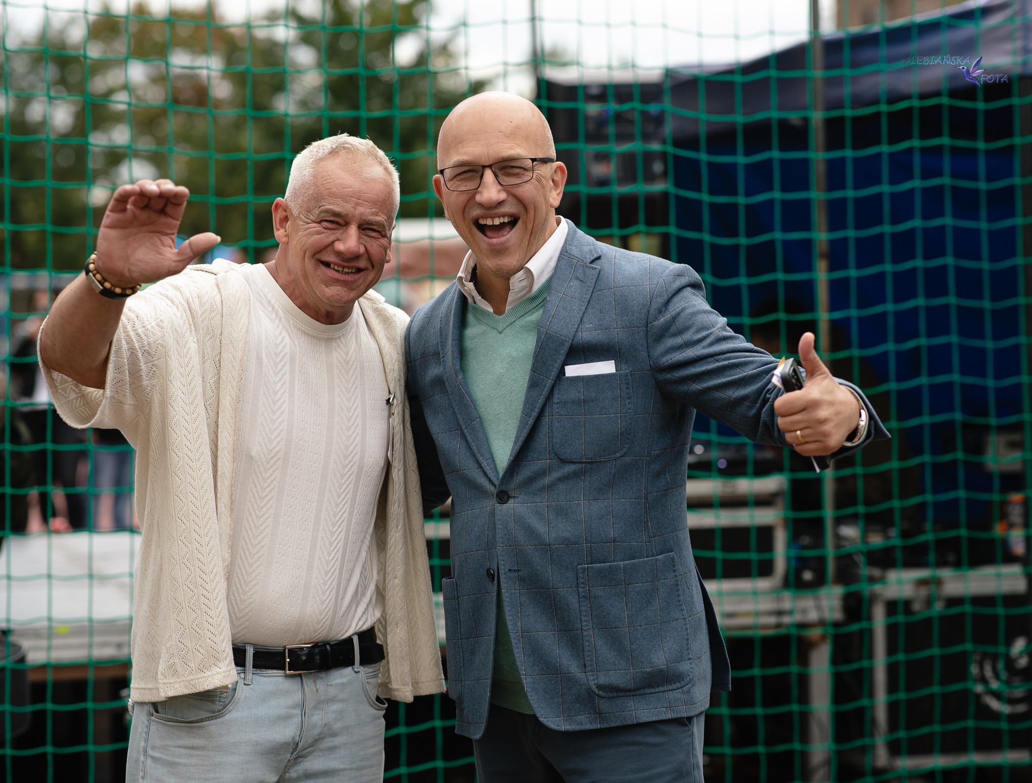 Uśmiechnięci mężczyźni pozują na tle siatki. Jeden w swetrze macha ręką, drugi w marynarce pokazuje kciuk w górę. Zdjęcie z eventu.