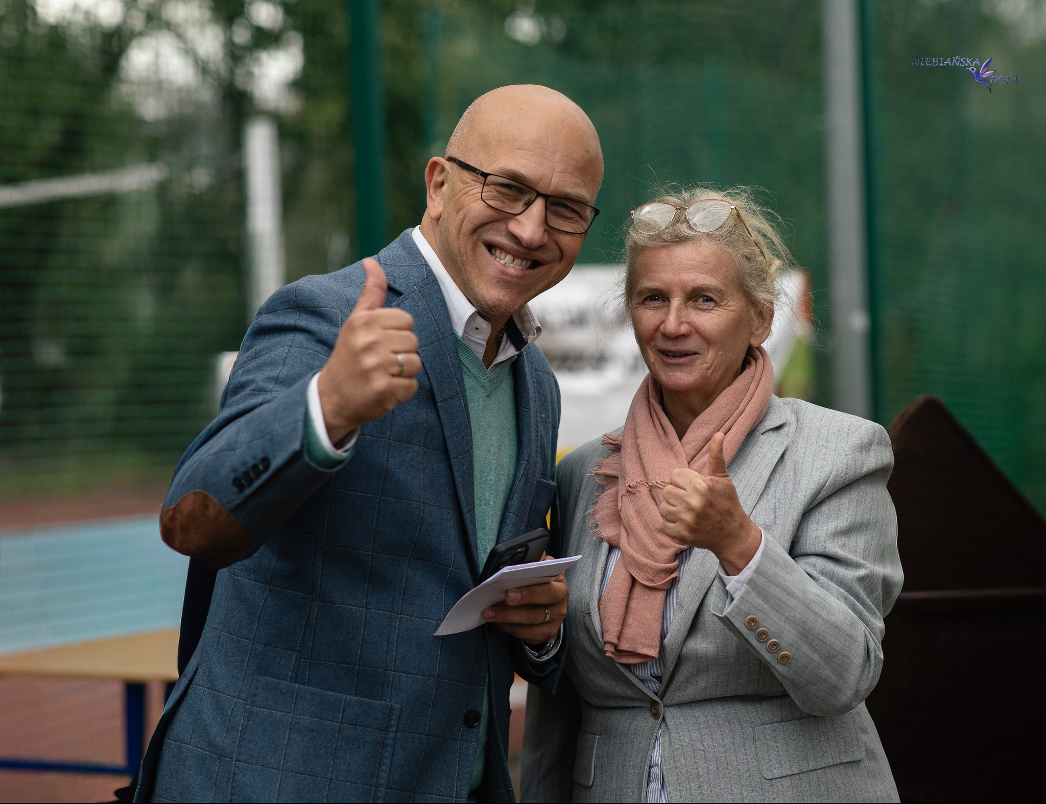 Portret uśmiechniętych osób w garniturach, pokazujących kciuki w górę na tle siatki ogrodzeniowej i stołu. Zdjęcie z lekkim cieniem i logo fotografa w prawym górnym rogu.