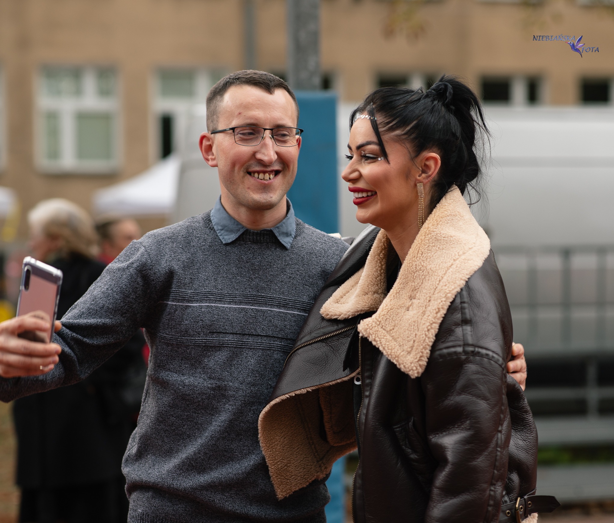 Uśmiechnięty mężczyzna robi selfie z kobietą w czarnej kurtce z futrzanym kołnierzem. Kobieta ma wyrazisty makijaż i ozdoby na twarzy. W tle budynek i fragmenty straganów.