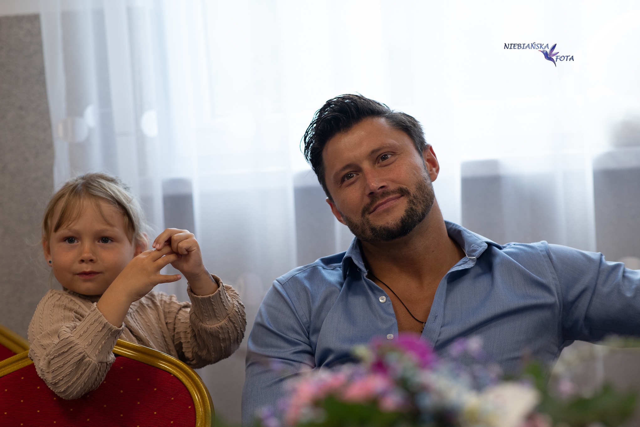 Portret uśmiechniętego mężczyzny w błękitnej koszuli obok małej dziewczynki z blond włosami, siedzących przy stole z kwiatami. W tle jasne zasłony.