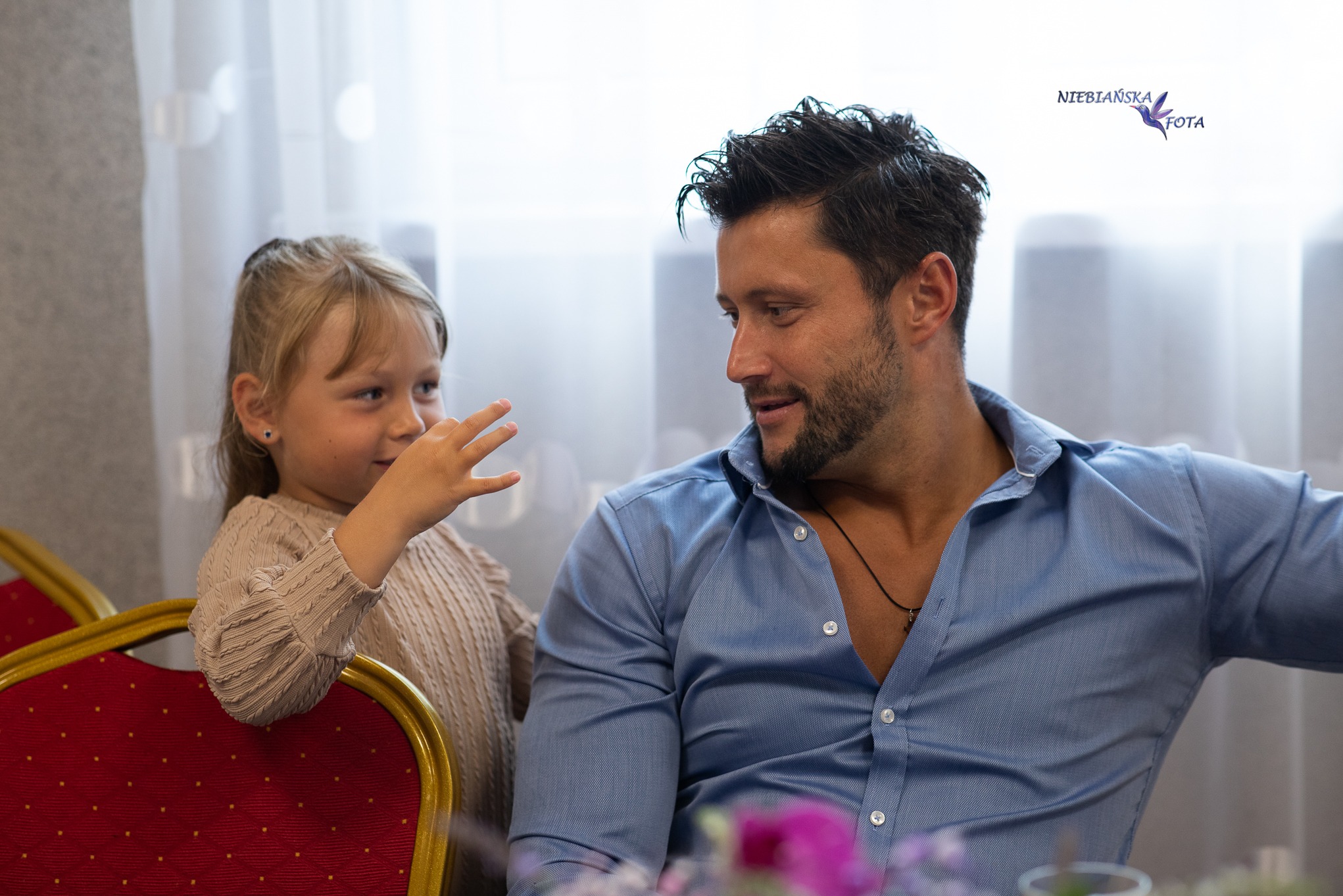Jasne, portretowe ujęcie ojca i córki; dziewczynka trzyma dłoń przed twarzą, oboje patrzą na siebie. W tle okno i czerwone krzesło z złotymi zdobieniami.