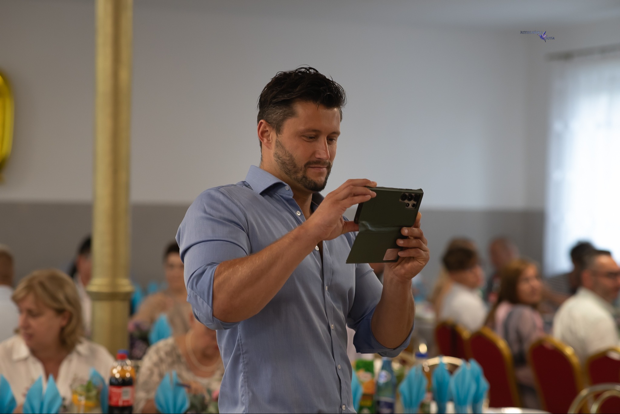 Mężczyzna w błękitnej koszuli robi zdjęcie telefonem na tle sali bankietowej z gośćmi, prawdopodobnie podczas imprezy okolicznościowej.