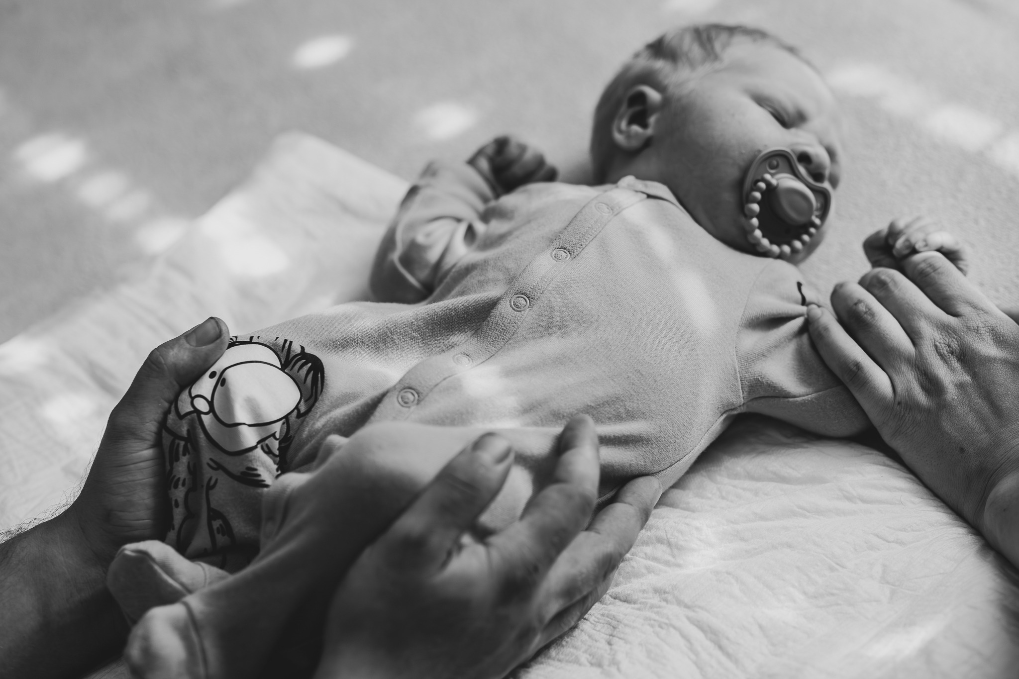 Czarno-białe zbliżenie śpiącego noworodka z smoczkiem, trzymanego w dłoniach rodziców na białym tle. Ubranko z motywem słonia.