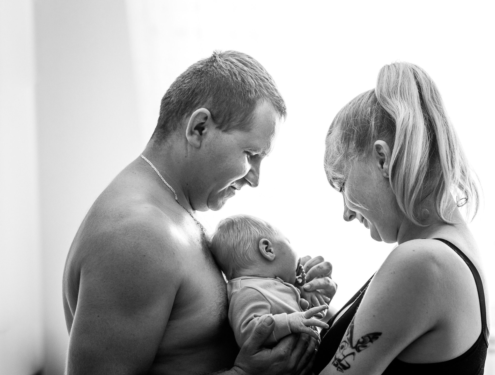 Czarno-białe zdjęcie rodziców spoglądających na niemowlę trzymane w ramionach ojca. Matka z tatuażem na ramieniu, ojciec bez koszuli, jasne tło.