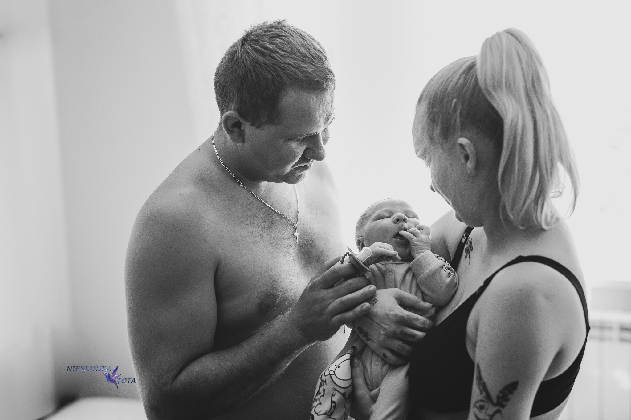 Czarno-białe zdjęcie rodziców z noworodkiem. Ojciec trzyma smoczek, matka przytula dziecko. Widoczne tatuaże na ciele matki. Minimalistyczne tło.