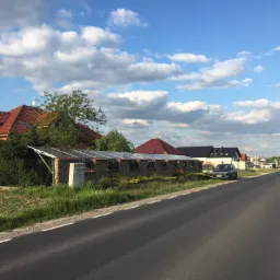 Instalacja paneli na konstrukcji przy posesji, widok od strony drogi, w tle domy jednorodzinne i niebo z chmurami.