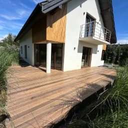 Nowoczesny dom z drewnianym tarasem otoczonym ozdobną trawą, widok z poziomu gruntu na taras i część elewacji z balkonem.