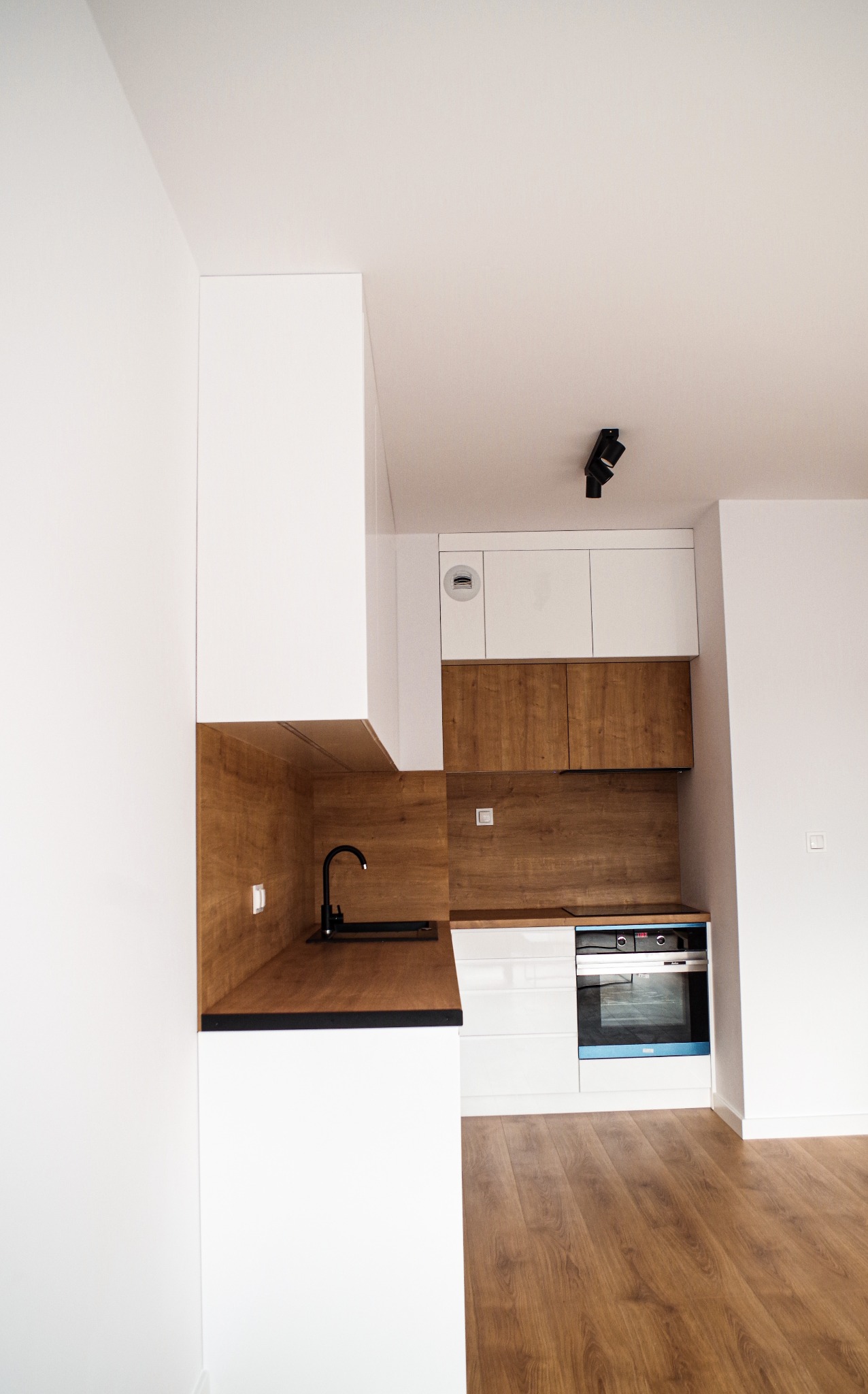 Nowoczesna kuchnia w stylu loftowym z białymi szafkami i drewnianymi akcentami, czarny zlew i armatura, oświetlenie punktowe na suficie, widok z perspektywy podłogi.