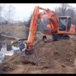 Pomarańczowa koparka Doosan na placu budowy, przygotowująca teren pod fundamenty, widoczne betonowe bloki fundamentowe i wykop ziemny, w tle drzewa.