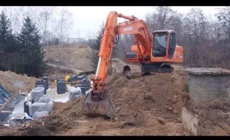 Pomarańczowa koparka Doosan na placu budowy, przygotowująca teren pod fundamenty, widoczne betonowe bloki fundamentowe i wykop ziemny, w tle drzewa.