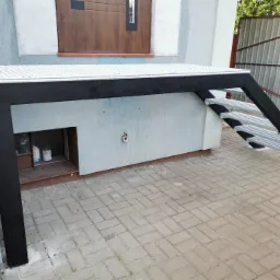 Metalowa konstrukcja schodów z podestem, prowadząca do drzwi, z kratowymi stopniami i czarną ramą, na tle elewacji budynku z małym okienkiem piwnicznym.