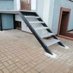 Zewnętrzne metalowe schody z ażurowymi stopniami prowadzące do drzwi, z widocznymi śladami po montażu na kostce brukowej.