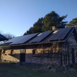 Ceglany budynek z panelami słonecznymi na dachu, widok z niskiej perspektywy, w tle drzewa i błękitne niebo.