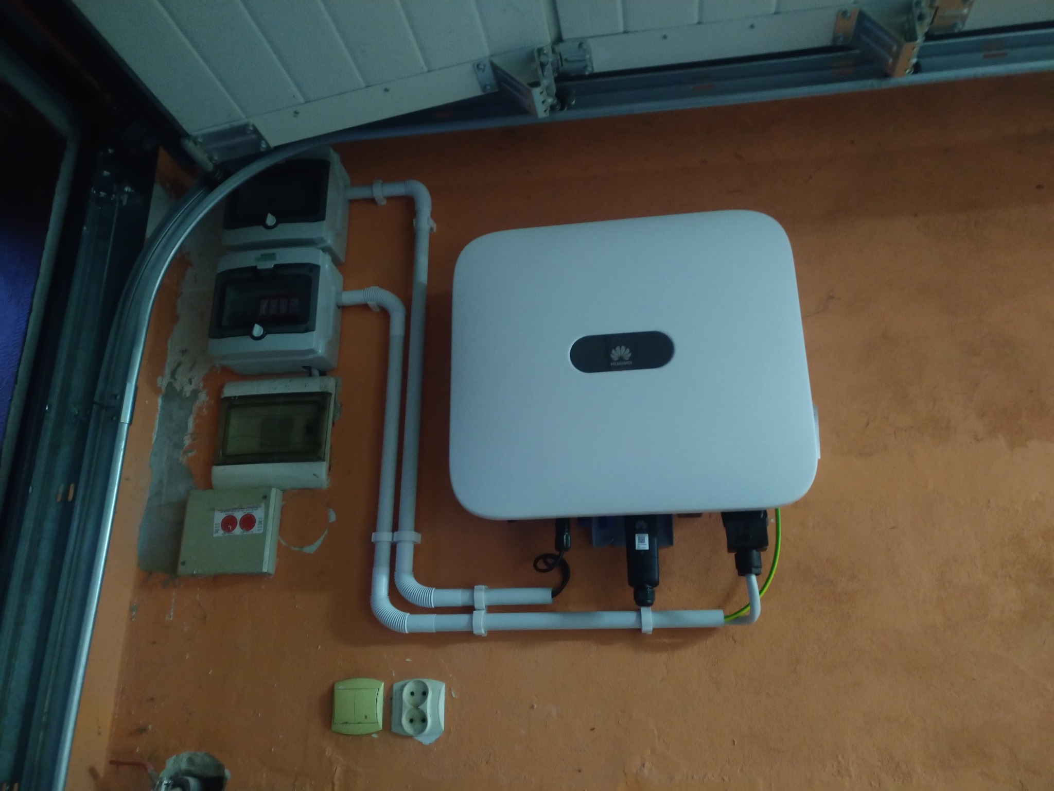 Biały falownik fotowoltaiczny zamontowany na pomarańczowej ścianie garażu, widoczne zabezpieczenia elektryczne i peszle instalacyjne.