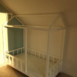 Biała drewniana rama łóżka dziecięcego w kształcie domku, z materacem, ustawiona w rogu pokoju z drewnianą podłogą i pomalowaną na biało-niebiesko ścianą.