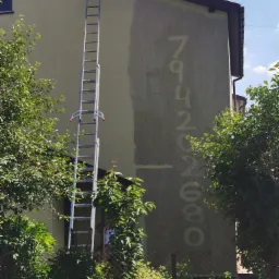 Fragment elewacji budynku z częściowo usuniętym numerem domu, widoczna drabina oparta o ścianę, otoczenie zieleni. Etap przygotowania ściany do malowania.