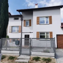 Dwukondygnacyjny dom z białymi ścianami, brązowymi okiennicami i szarą, poziomą bramą w tle, widziany zza szarego, metalowego ogrodzenia.