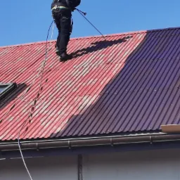 Częściowo umyty blaszany dach z widocznym oknem dachowym i osobą w uprzęży na dachu, z której spuszczony jest przewód.