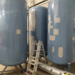 Trzy duże, niebieskie zbiorniki przemysłowe z widocznymi łatami szpachli, przygotowywane do malowania, z aluminiową drabiną przystawioną do środkowego zbiornika i rurą na podłodze.