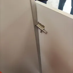 Zasuwa zabezpieczająca na drzwiach kabiny w toalecie publicznej; widoczne fragmenty osób w tle.