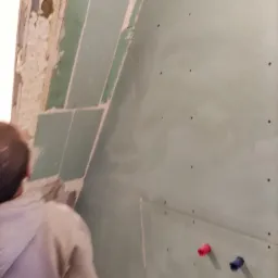 Montaż płyt karton gips na ścianie w łazience, widoczne fragmenty starej ściany i wystające rury instalacji wodnej z czerwonym i niebieskim oznaczeniem.