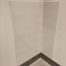 Świeżo ułożone, białe, prostokątne płytki na ścianie w rogu łazienki, widoczna ciemna fuga przy podłodze i wiaderko z zaprawą w dolnym rogu.