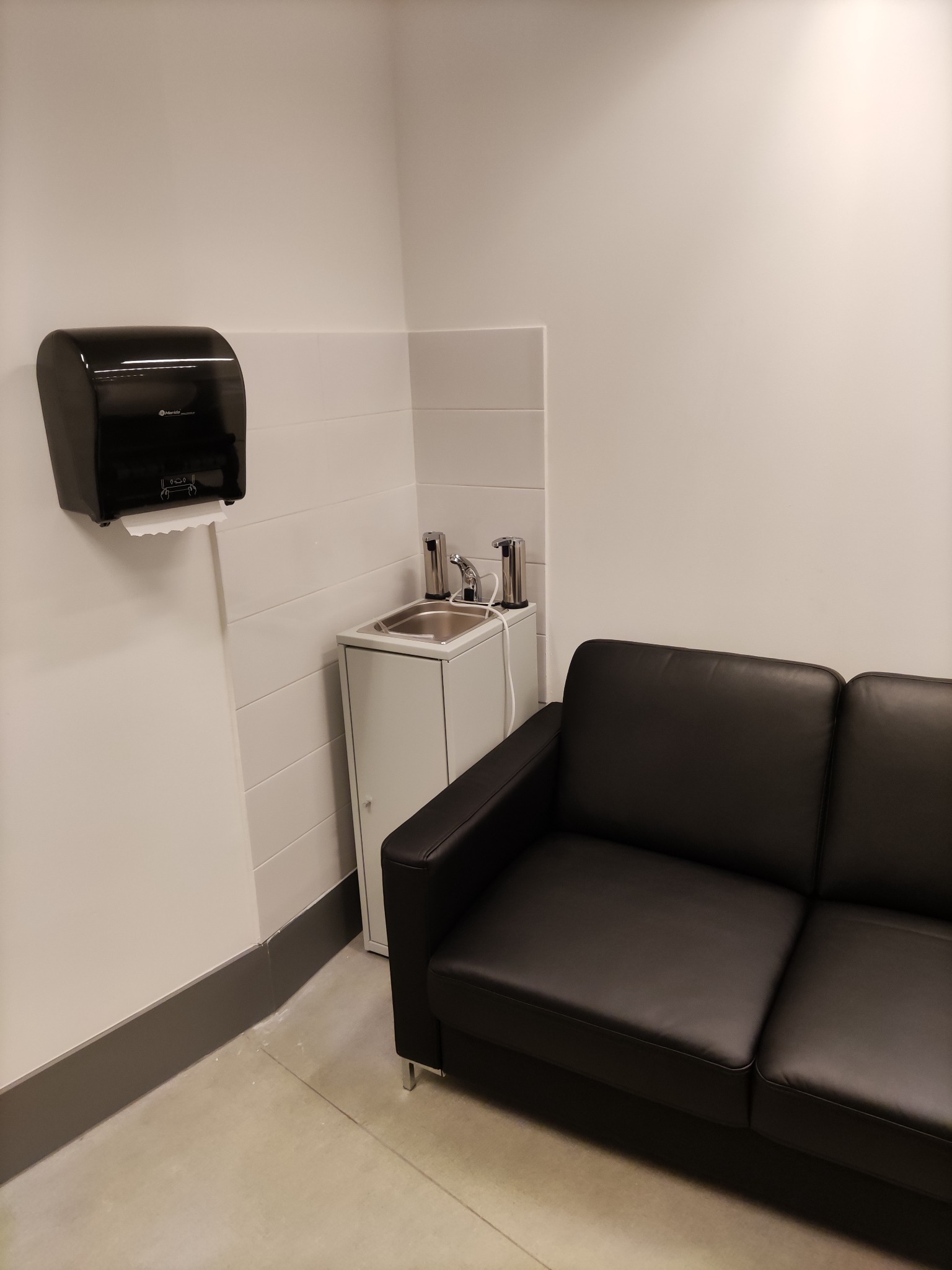 Minimalistyczna poczekalnia z czarną sofą, umywalką z dozownikiem i podajnikiem ręczników papierowych na białej ścianie z szarą listwą przypodłogową.