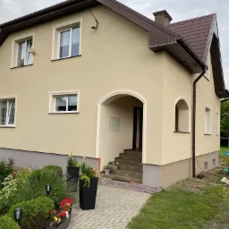 Beżowy dom jednorodzinny z brązowym dachem, widoczny z boku, z zadbanym ogrodem, podjazd z kostki, widoczne elementy instalacji na elewacji.