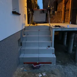 Schody zewnętrzne wyłożone jasnymi płytkami w trakcie prac wykończeniowych, widoczny poziomica, pojemnik z akcesoriami do układania płytek, elewacja z szarego tynku strukturalnego.