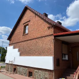 Ceglany dom w trakcie ocieplania: białe płyty izolacyjne częściowo zamontowane na ścianie, drabina oparta o ścianę, widoczne niebo i chmury.