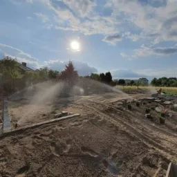 Instalacja automatycznego nawadniania trawnika w trakcie zakładania ogrodu, widoczne zraszacze w akcji, przygotowane rośliny w doniczkach i obrzeża z kostki.