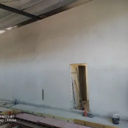 Świeżo otynkowana ściana w surowym wnętrzu hali, widoczny pracownik w czerwonej czapce, przygotowujący się do dalszych prac wykończeniowych, z widocznymi śladami po szalunkach na podłodze.