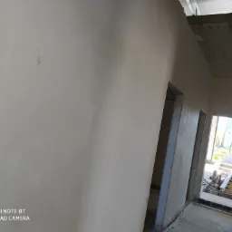 Ściana wewnętrzna po nałożeniu wstępnej warstwy, widoczne niedoskonałości i ślady po szpachlowaniu, surowy stan wykończenia z widocznymi ramami drzwiowymi i konstrukcją stropu.