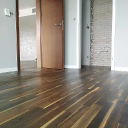 Wypolerowana, ciemna drewniana podłoga w nowym wnętrzu z otwartymi drzwiami do łazienki i pokoju z dekoracyjną ścianą.