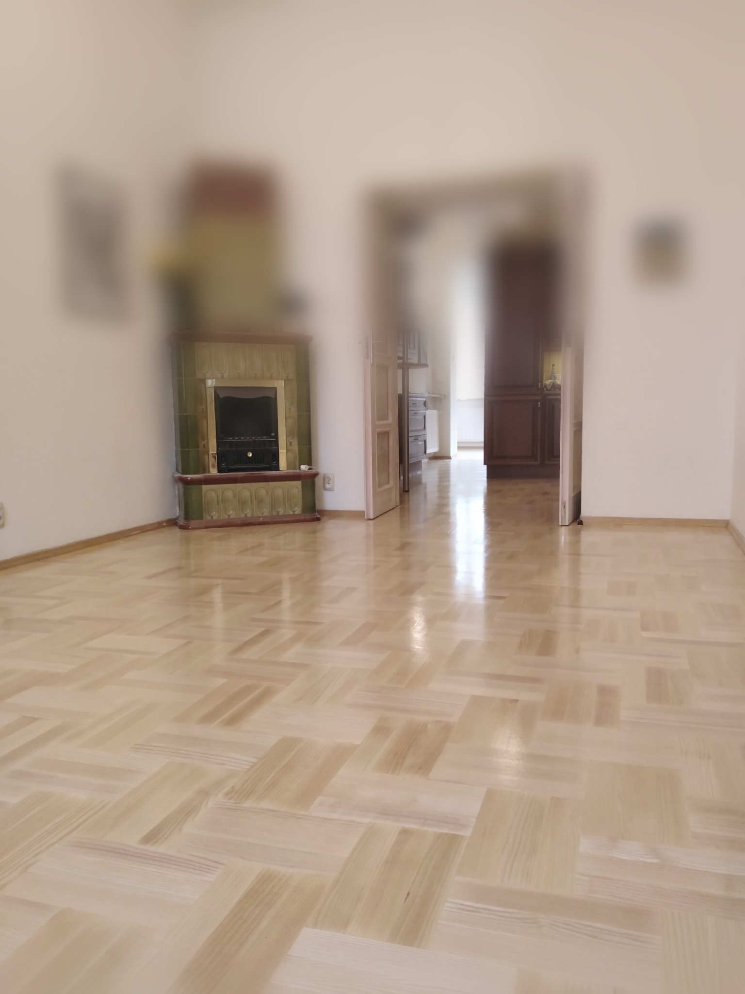 Jasna, odnowiona parkietowa podłoga w pokoju z zielonym, kaflowym piecem i otwartymi drzwiami do dalszej części mieszkania.
