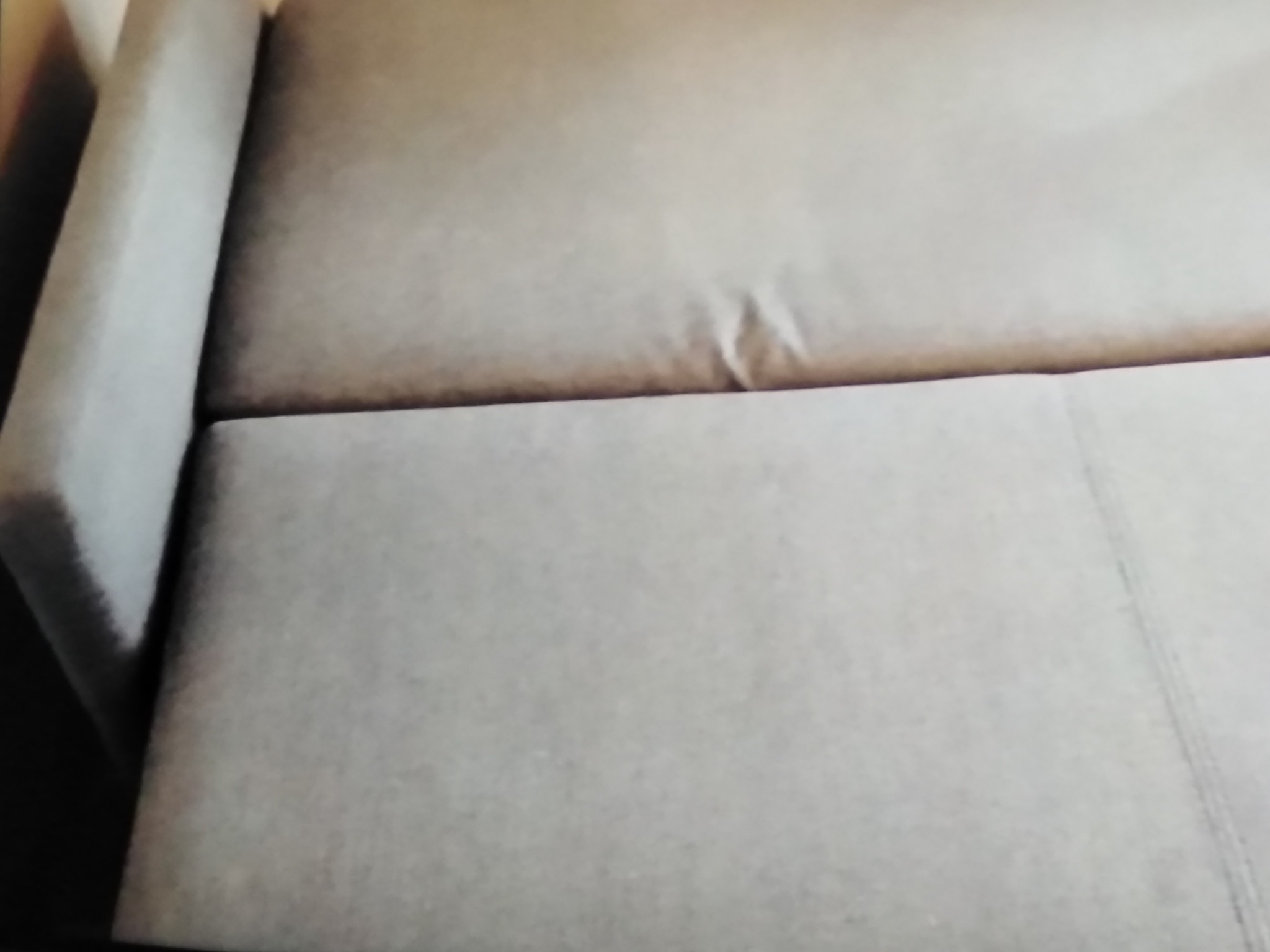 Fragment jasnoszarej kanapy z widocznymi załamaniami na materiale i delikatnym przeszyciem wzdłuż siedziska.