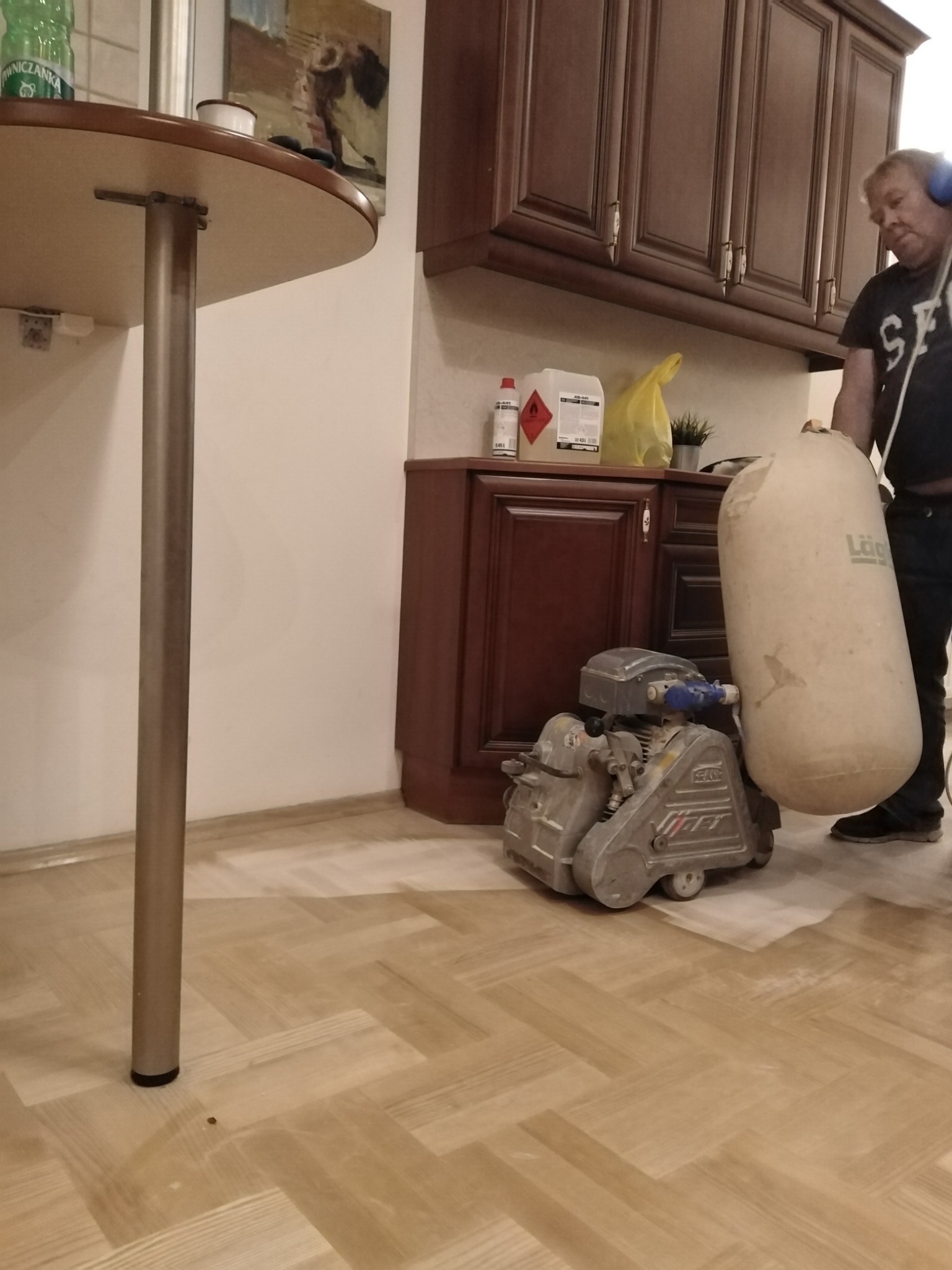 Pracownik w słuchawkach obsługuje maszynę do renowacji parkietu, widoczny fragment odnowionej podłogi w jodełkę, w tle meble kuchenne.