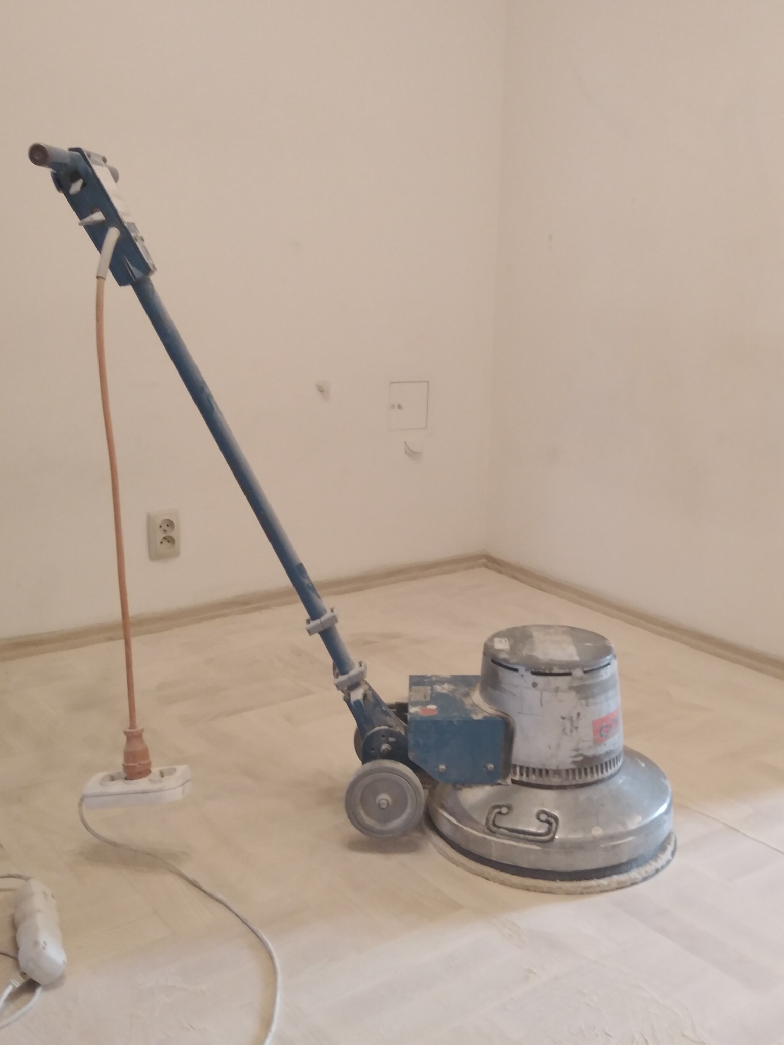 Maszyna do renowacji podłóg parkietowych z podłączonym kablem zasilającym w trakcie pracy, widoczne ślady pyłu po szlifowaniu.