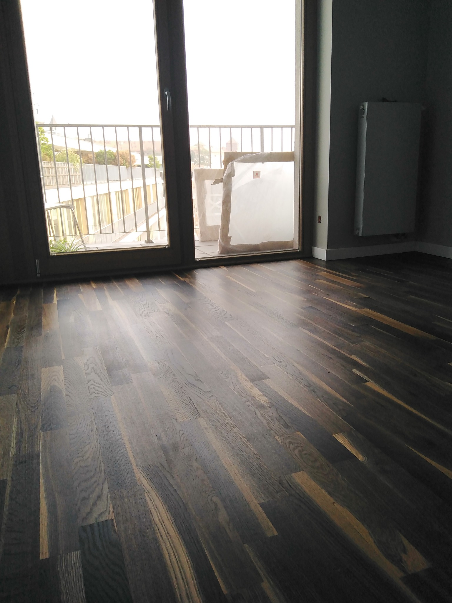 Wypolerowany parkiet w odcieniach brązu i szarości w pustym pokoju z balkonem i grzejnikiem na ścianie.