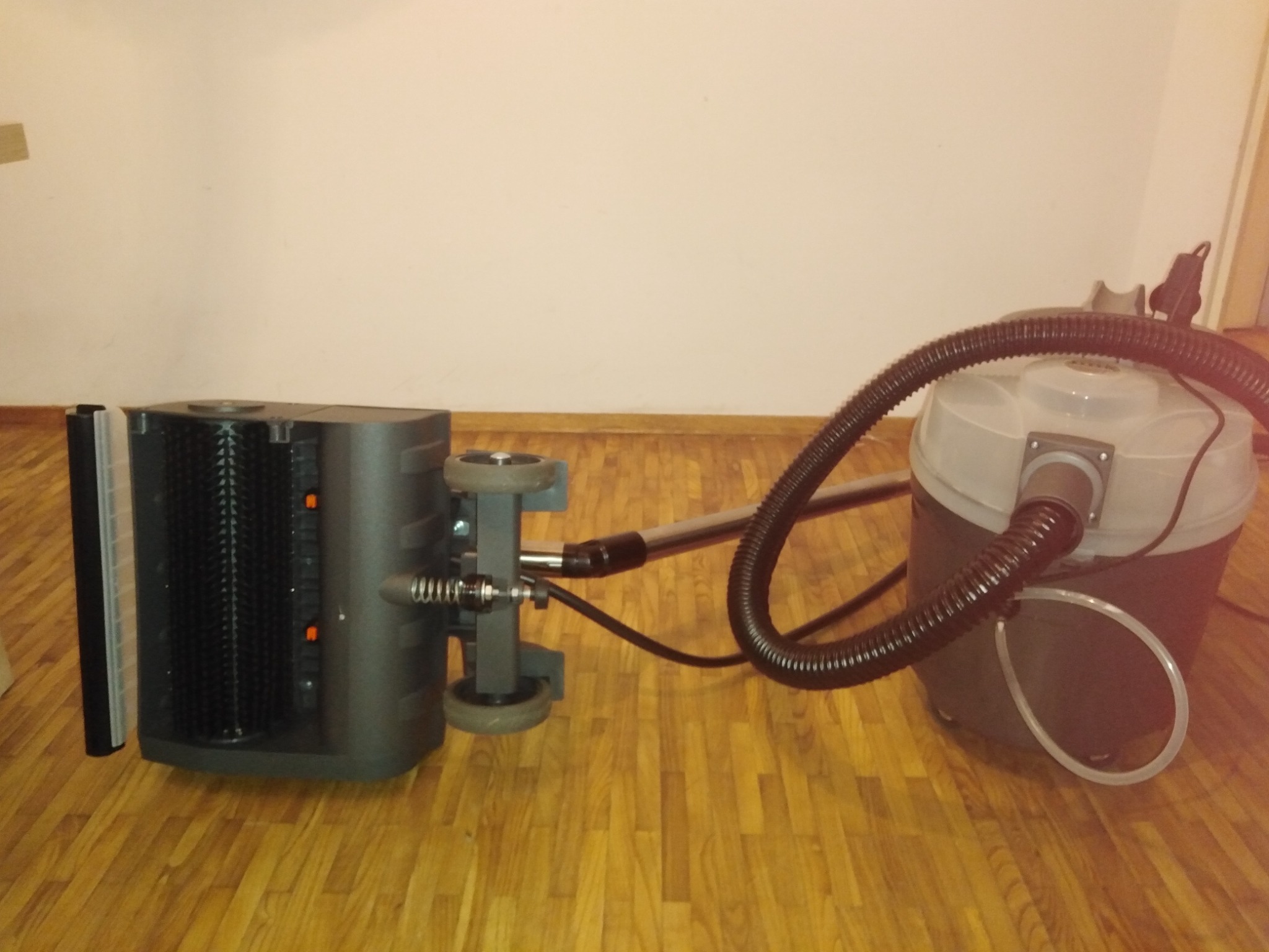 Parkieciarka połączona wężem z odkurzaczem przemysłowym, stojąca na drewnianej podłodze z widoczną ścianą w tle.