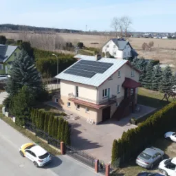 Dom jednorodzinny z panelami słonecznymi na dachu, widok z góry na posesję z ogrodzeniem i zaparkowanymi samochodami, w tle zabudowa wiejska.