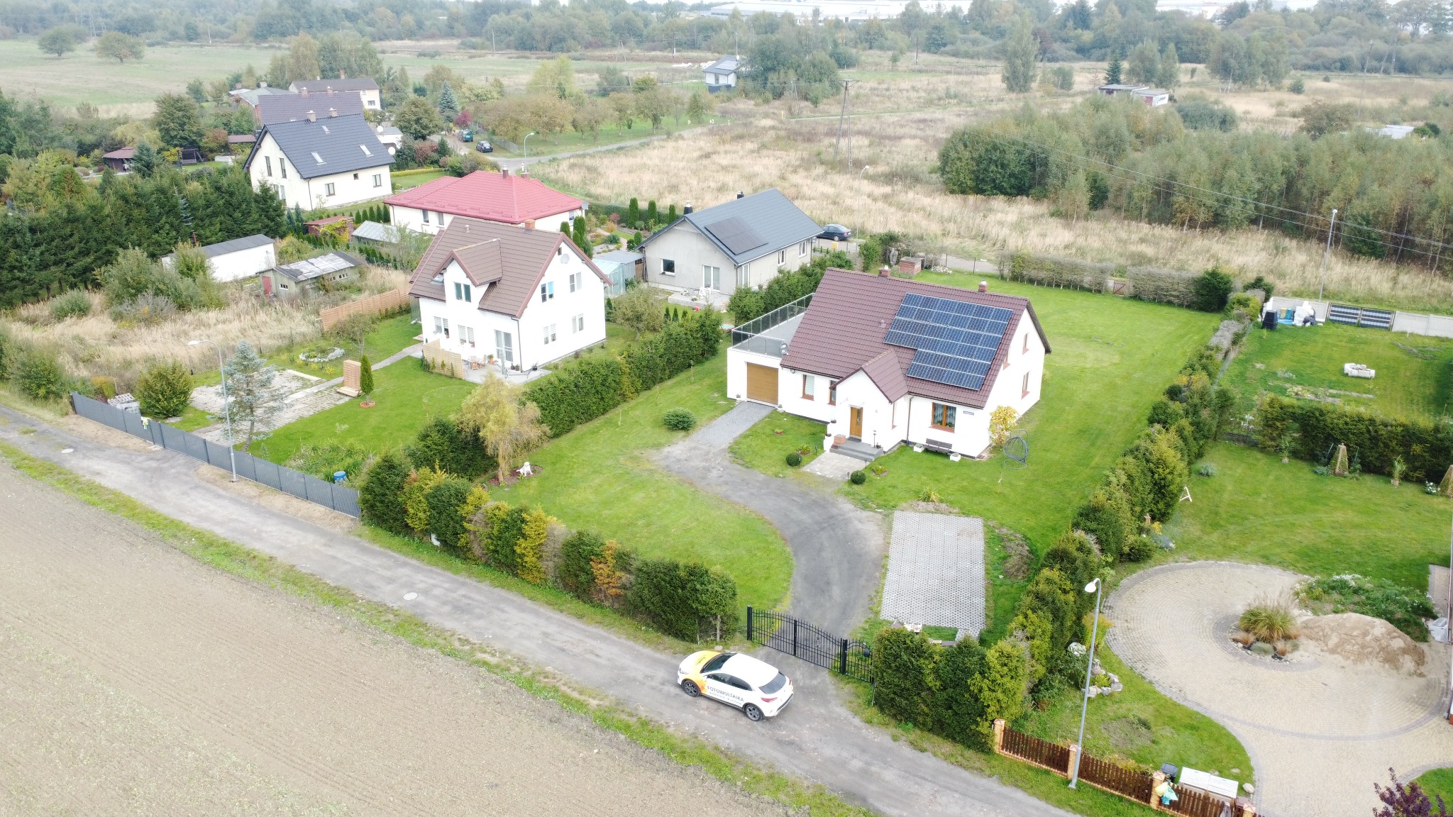 Widok z drona na dom jednorodzinny z panelami słonecznymi na dachu, biały samochód z żółtym akcentem zaparkowany przed posesją, zadbany ogród i inne domy w tle.