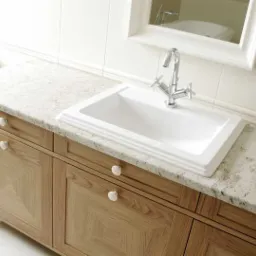 Biała, prostokątna umywalka z chromowaną baterią na blacie z jasnego granitu, osadzona na szafce z drewnianymi frontami o geometrycznym wzorze, uzupełniona okrągłymi, białymi uchwytami.