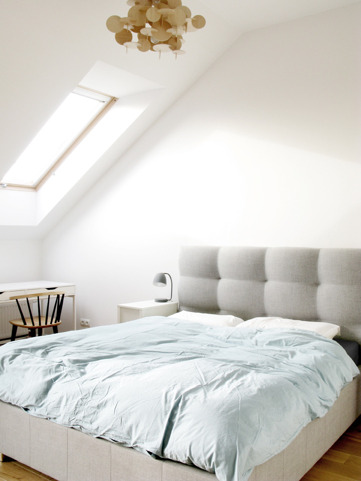 Jasne poddasze w Krakowie z łóżkiem z szarą tapicerką, drewnianą lampą wiszącą i oknem dachowym w skosie.